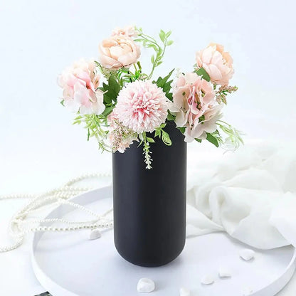 Black Cylinder Vase on a white table with pink flowers inside and white decorative elements next to it