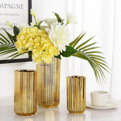 Three Cylinder Gold Vases with flowers inside on a white table next to a mug