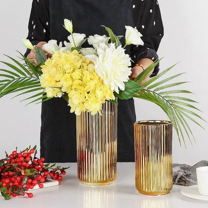 Two Cylinder Gold Vases with yellow flowers inside on a white shiny table