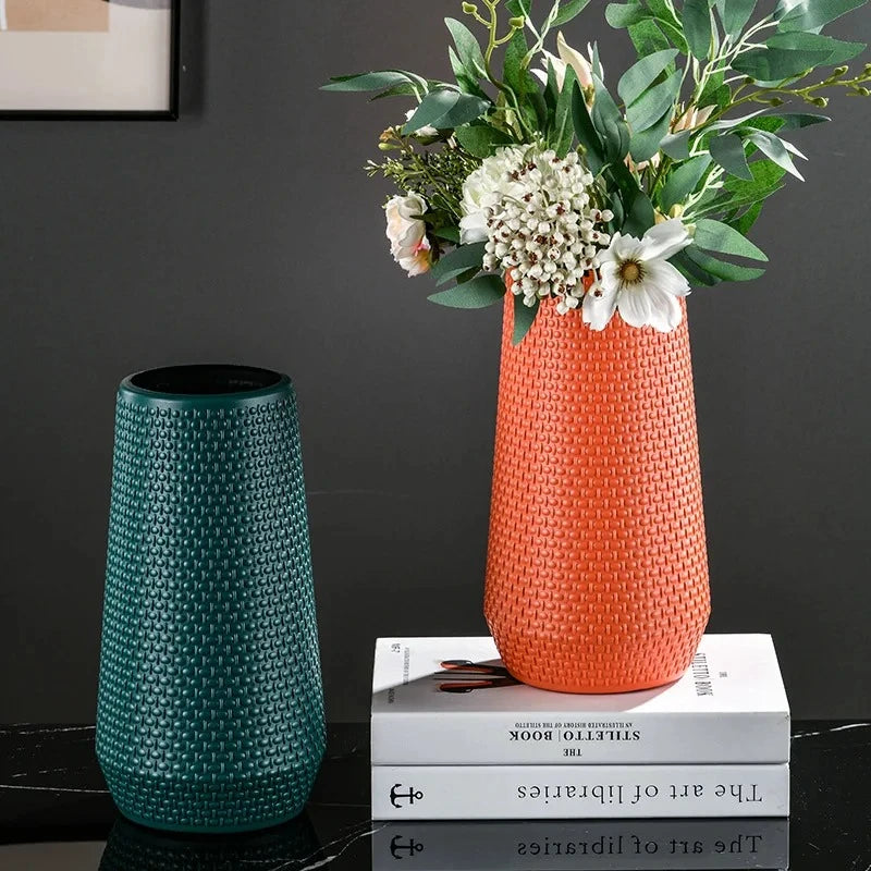 Two Plastic Cylinder Vases in teal and orange with flowers inside standing on a book